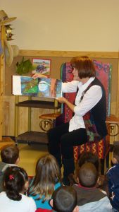 A female instructors is shown sitting in a chairwoman and reading ampere picture book to a group of children sitting by front on her off the store.